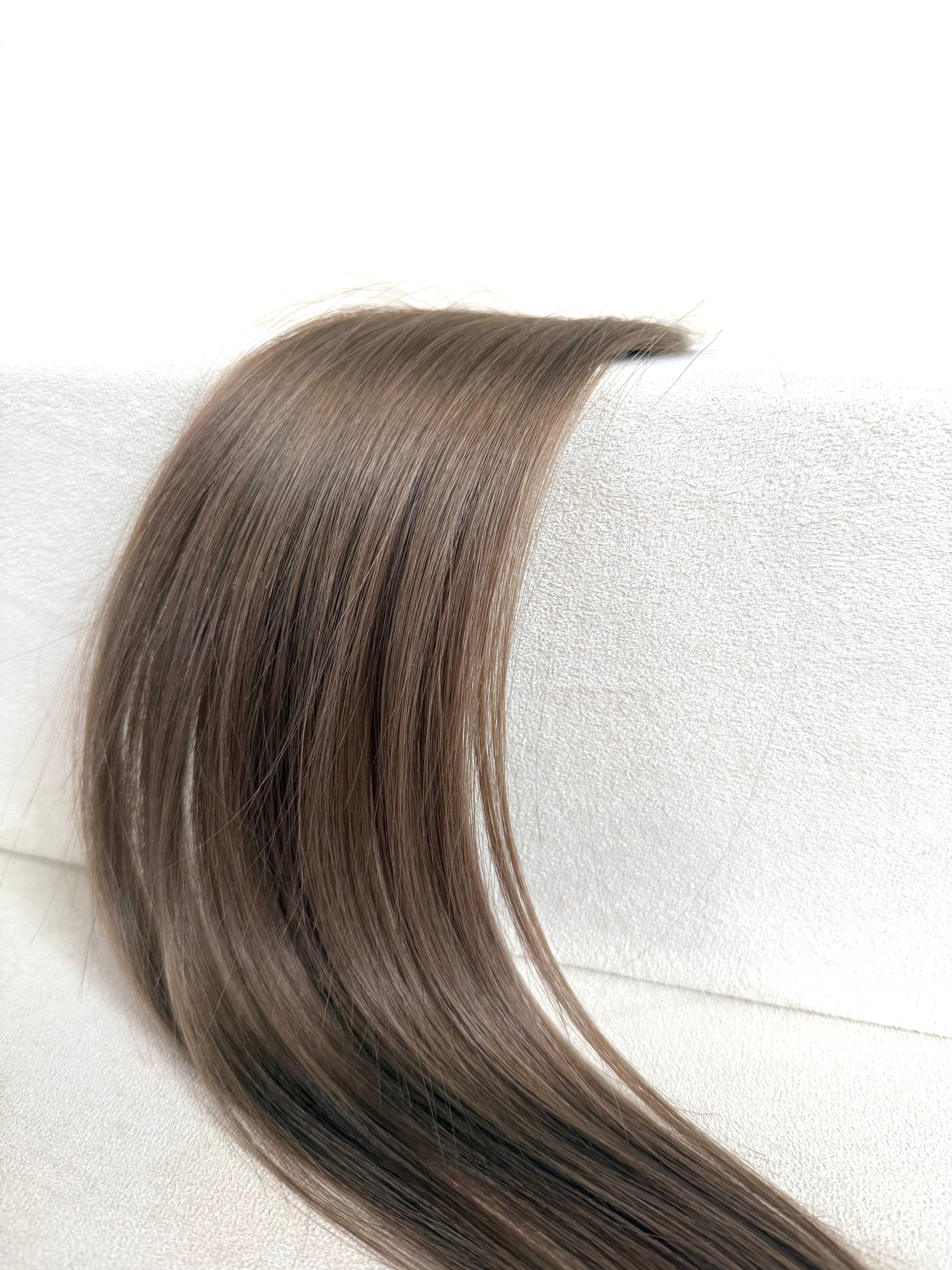 A long, straight hair extension or wig section in a medium brown shade, draped over a white textured surface against a white background.